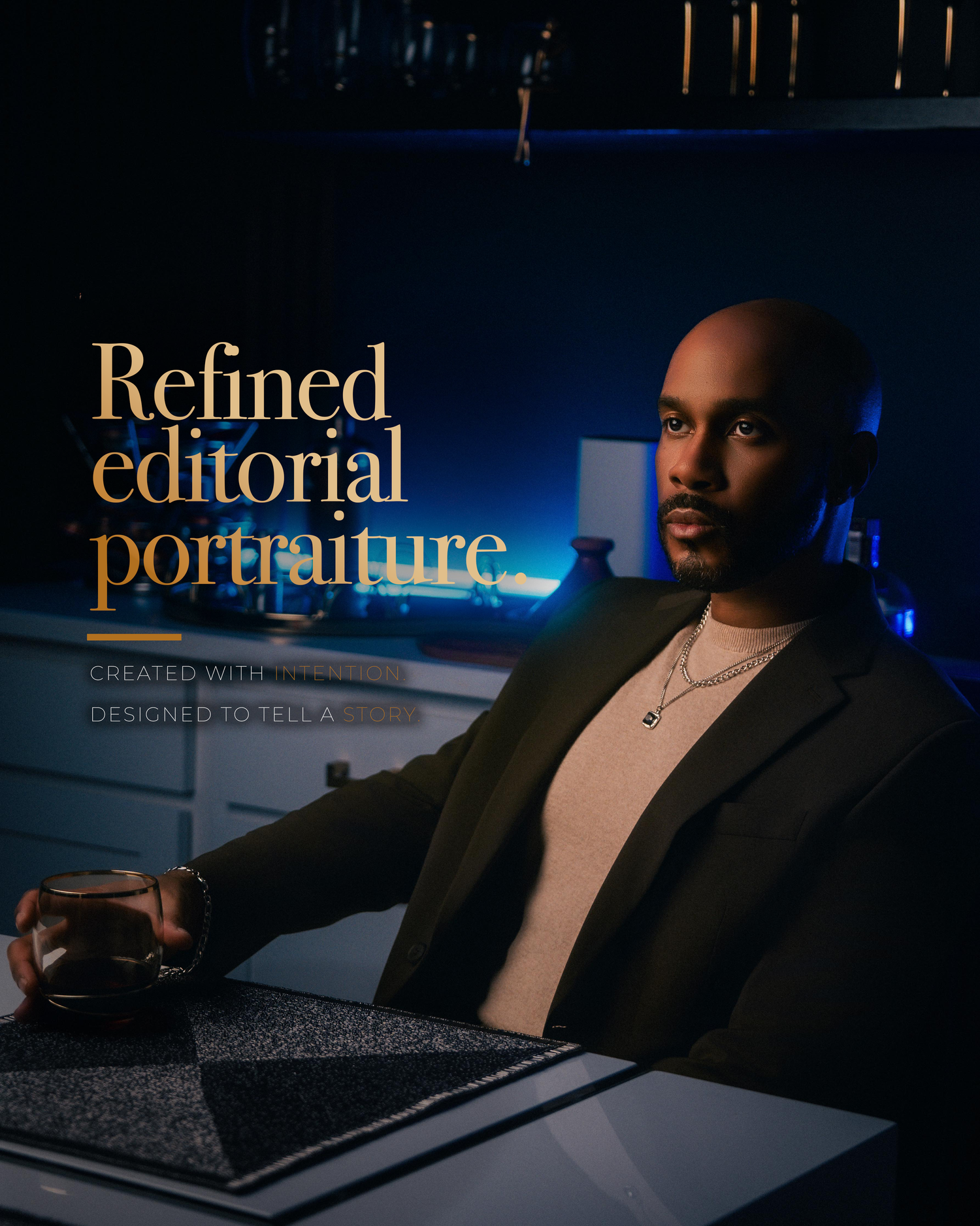 A bald Black man in a dark blazer sits confidently at a desk holding a glass in moody blue studio lighting.