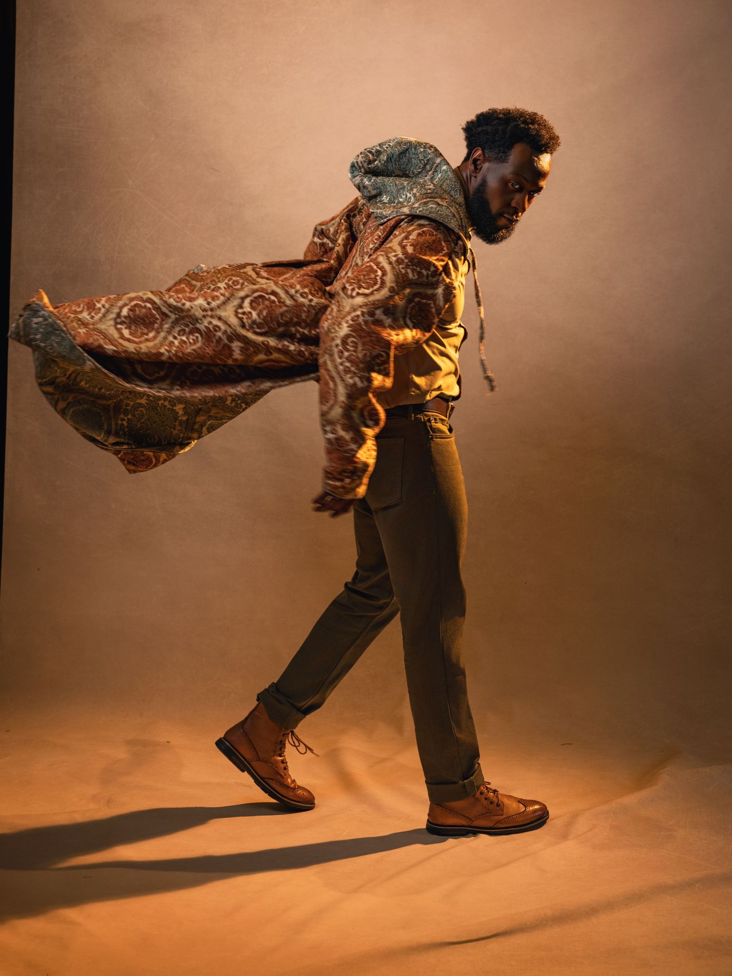 Man walking confidently in patterned cape, yellow hoodie, olive pants, and brown boots against warm studio backdrop in cinematic portrait