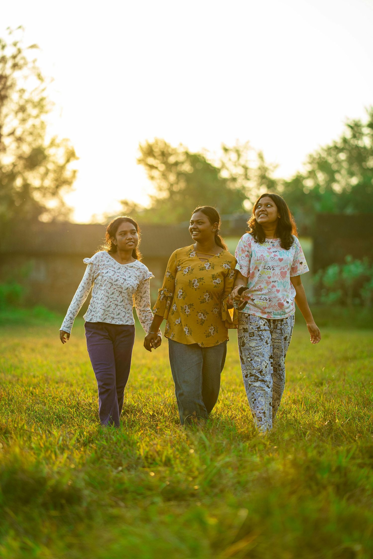Three women walking hand-in-hand through a sunlit green field at golden hour, smiling and enjoying time together outdoors.