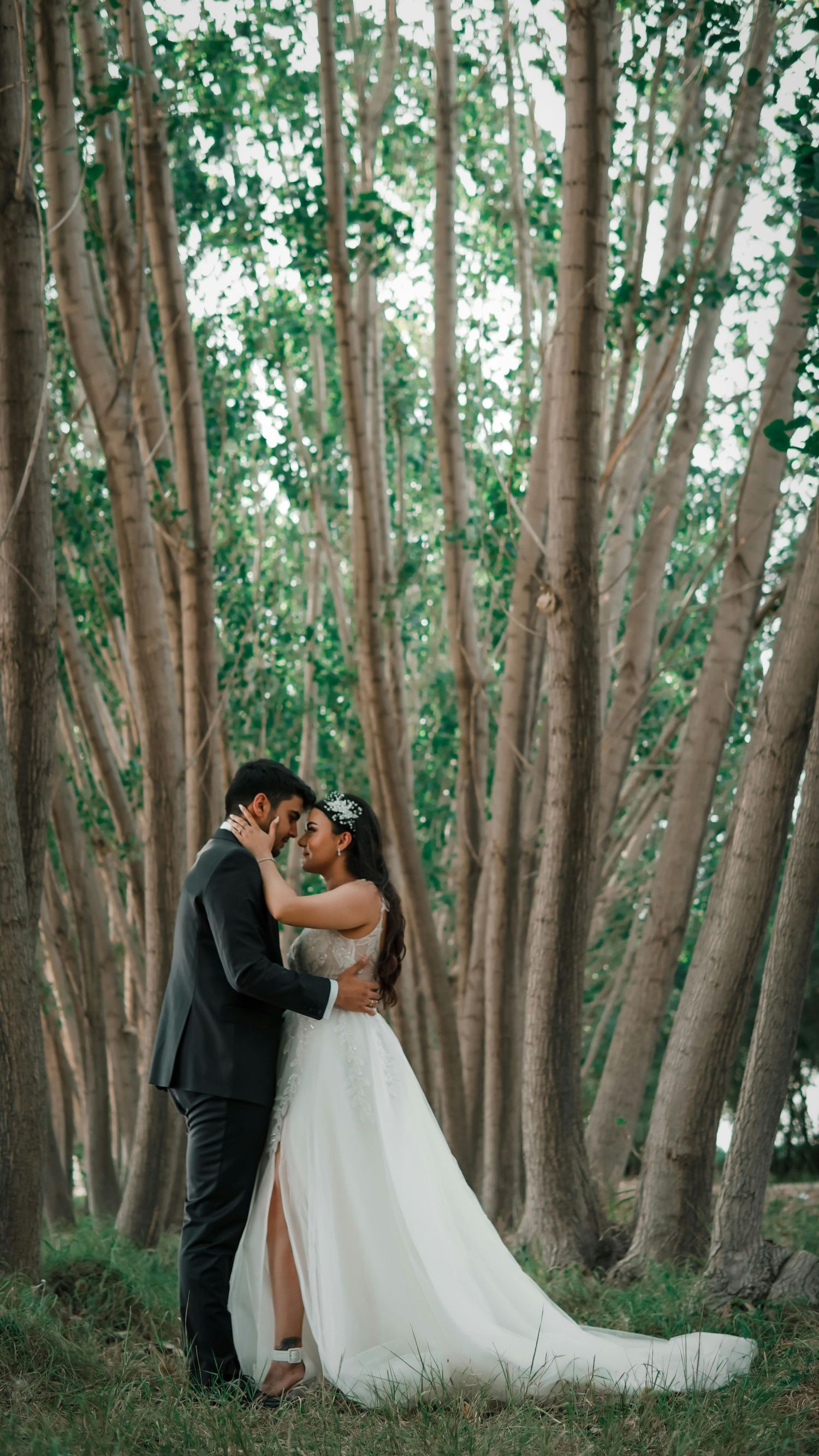 A bride and groom share a romantic embrace in a lush forest setting, surrounded by tall trees.