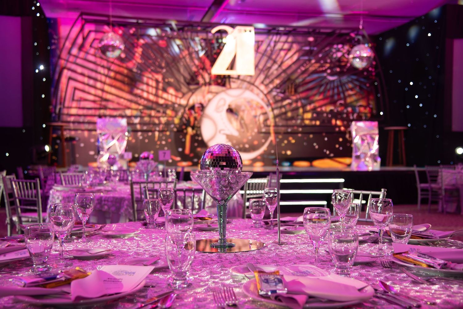 Elegant banquet hall with pink lighting, crystal glassware, and disco ball centerpieces set for a glamorous gala event.