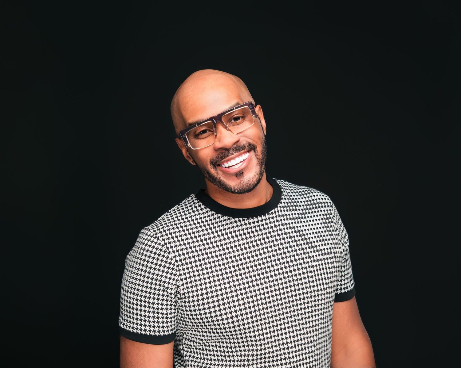 A smiling bald man with glasses wearing a houndstooth t-shirt against a dark background captured by Atlanta lifestyle photographer