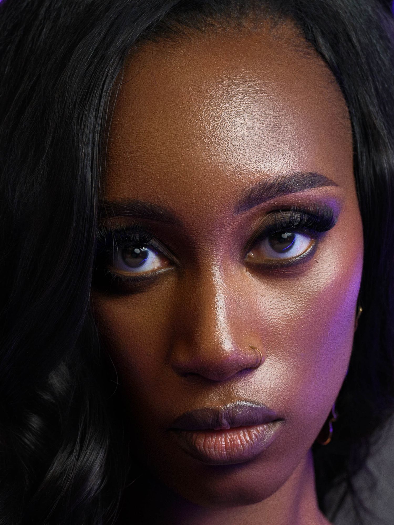 Close-up portrait of a young Black woman with glamorous purple eye makeup, long lashes, and dark lips under moody lighting.