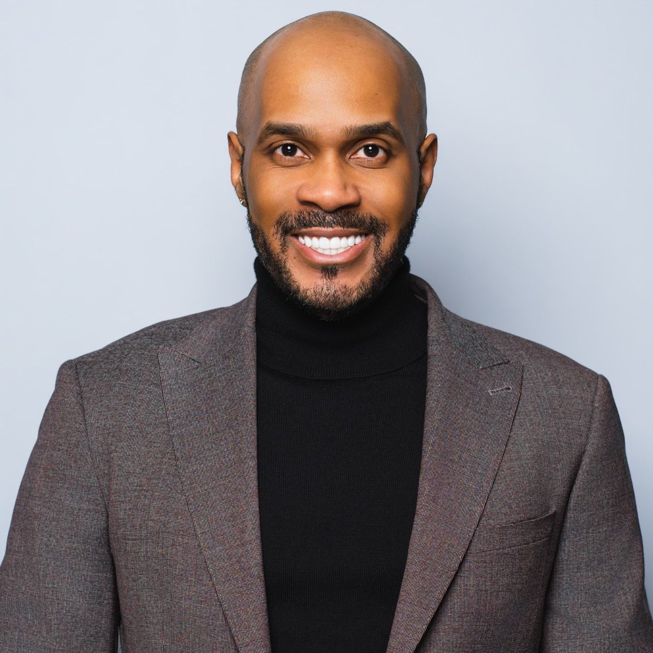 Professional headshot of a bald Black man in gray blazer and black turtleneck smiling confidently.