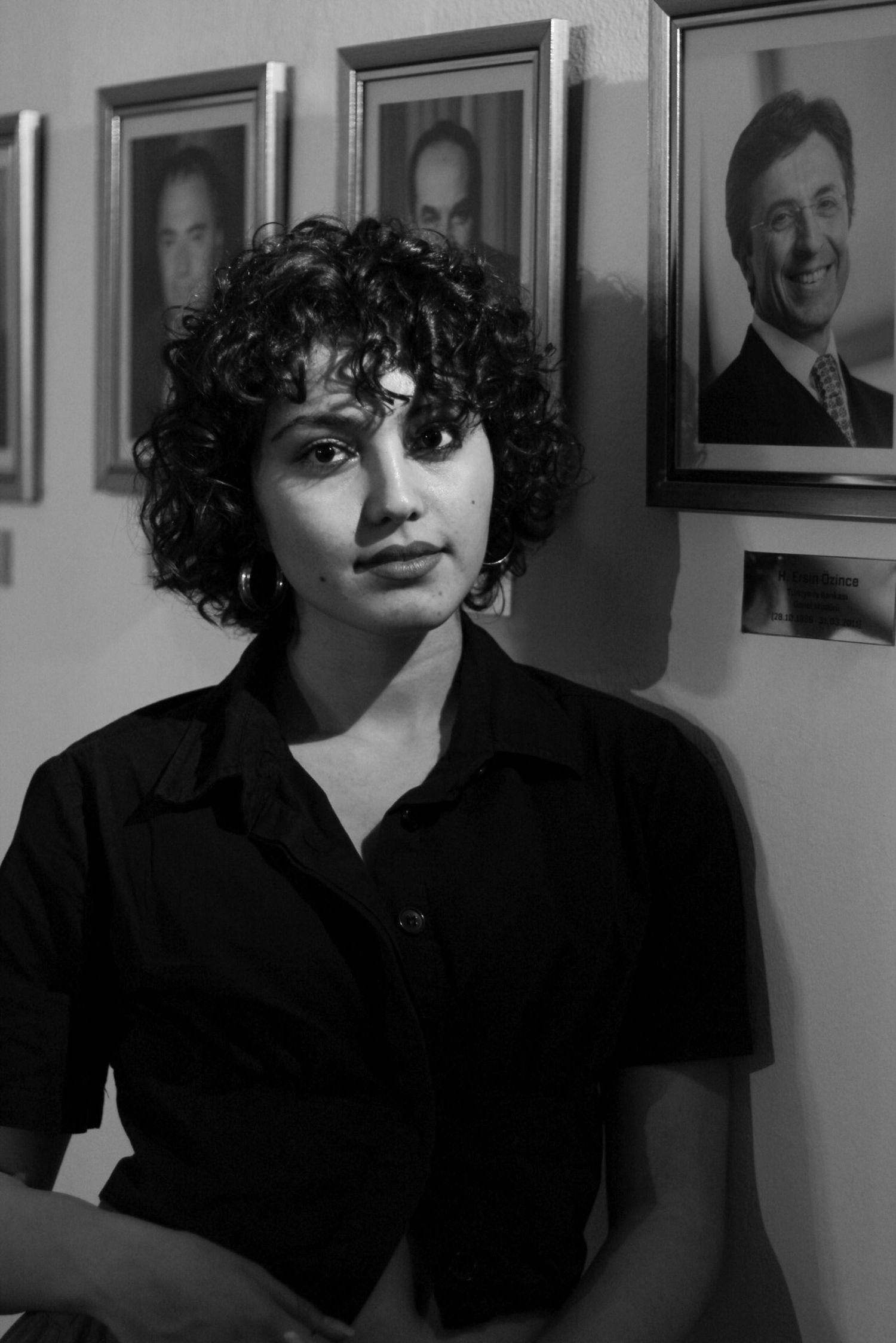 Black and white portrait of a young woman with curly hair in black shirt, framed photos on wall behind her.