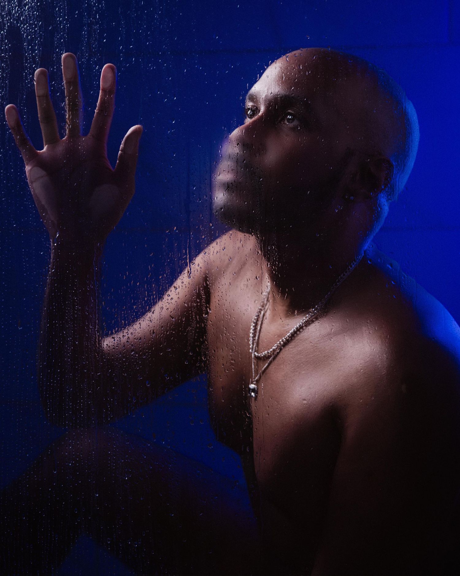 Shirtless bald man with chain necklace pressing hand against wet glass with blue dramatic lighting captured by ATL cinematic photographer