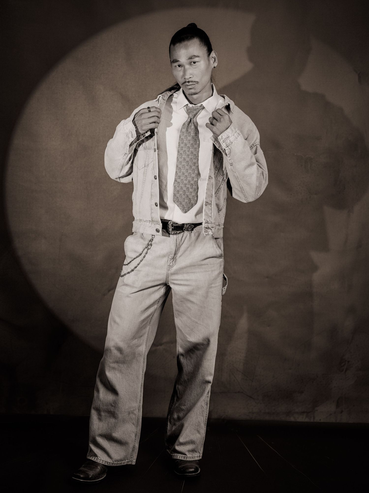 Black and white portrait of a man in wide-leg pants, jacket, and tie posing confidently in a studio.