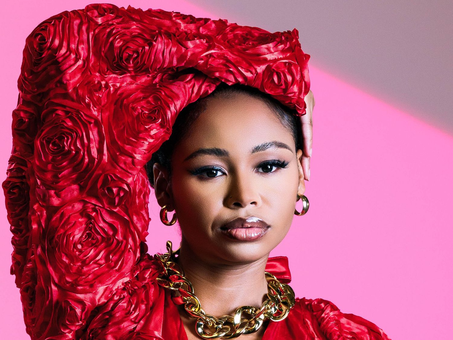Woman in bold red floral outfit and gold chain necklace poses against a pink background.