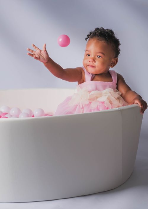 A joyful baby girl in a pink dress plays in a white ball pit, tossing a pink ball with a bright smile by ATL photographer
