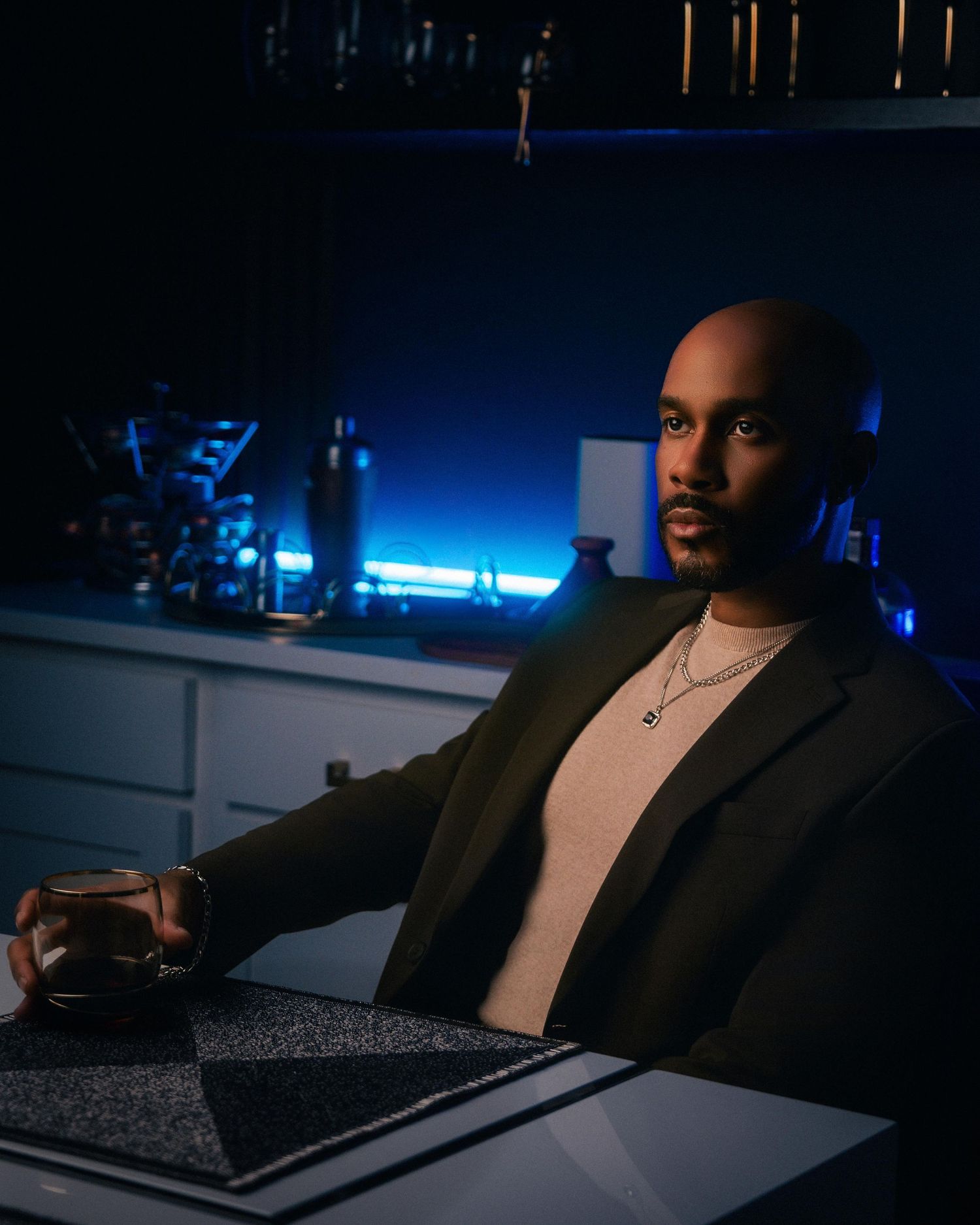 Stylish bald man in dark blazer holding whiskey glass at bar with blue neon lighting backdrop.