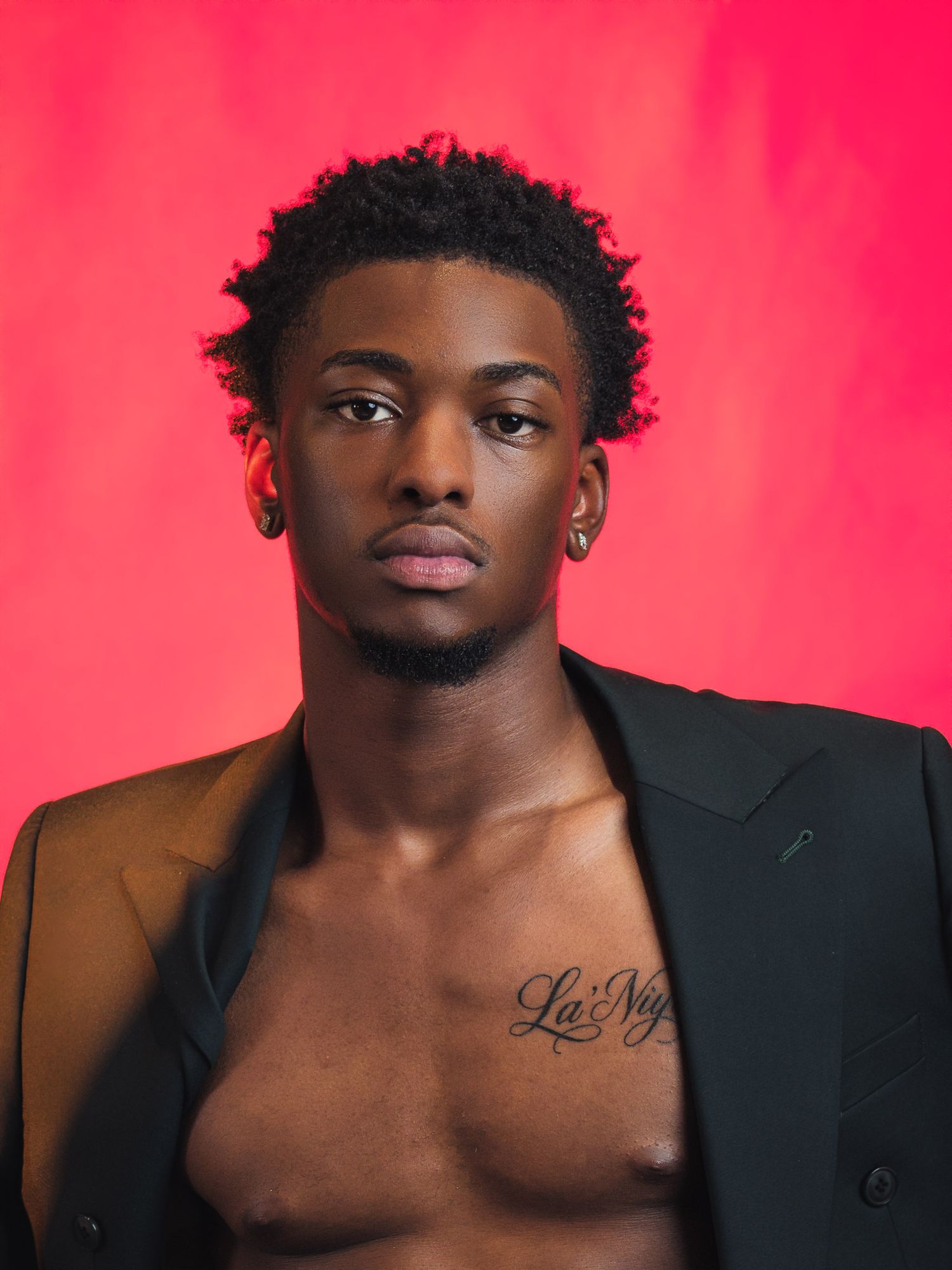 Young man in open black blazer with chest tattoo posing against vibrant red background.