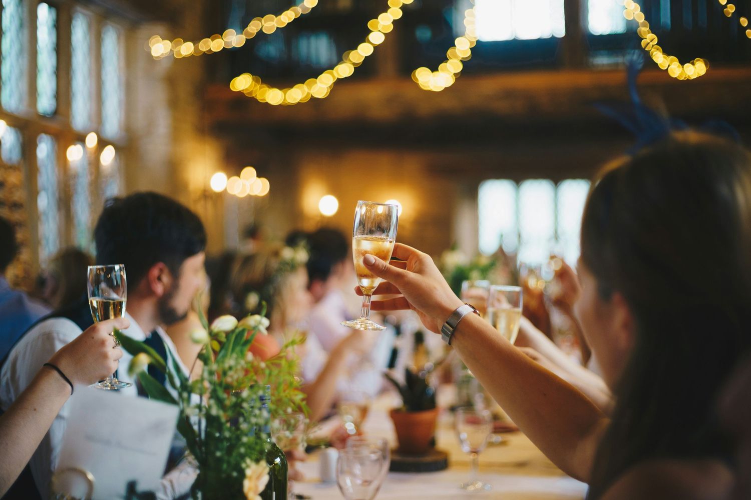 Guests raising champagne glasses in a toast at an elegant wedding reception with warm string lights.