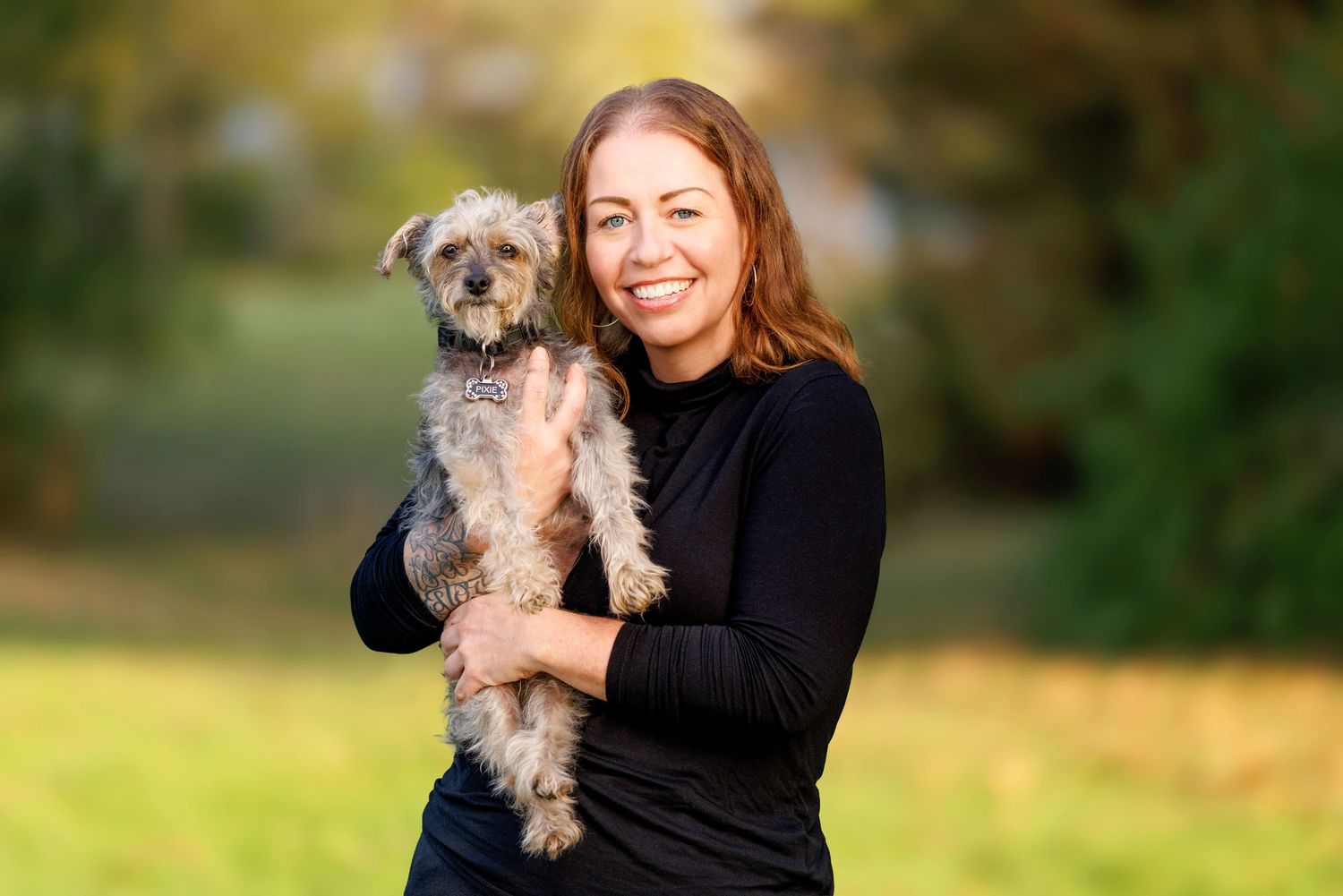 A smiling woman in black holds a small gray terrier dog outdoors in a park setting.