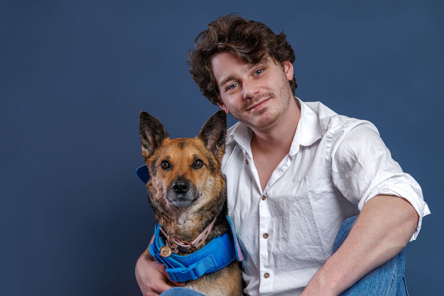 A young man with curly hair in a white shirt hugs a German Shepherd dog wearing a blue vest against a dark blue background.