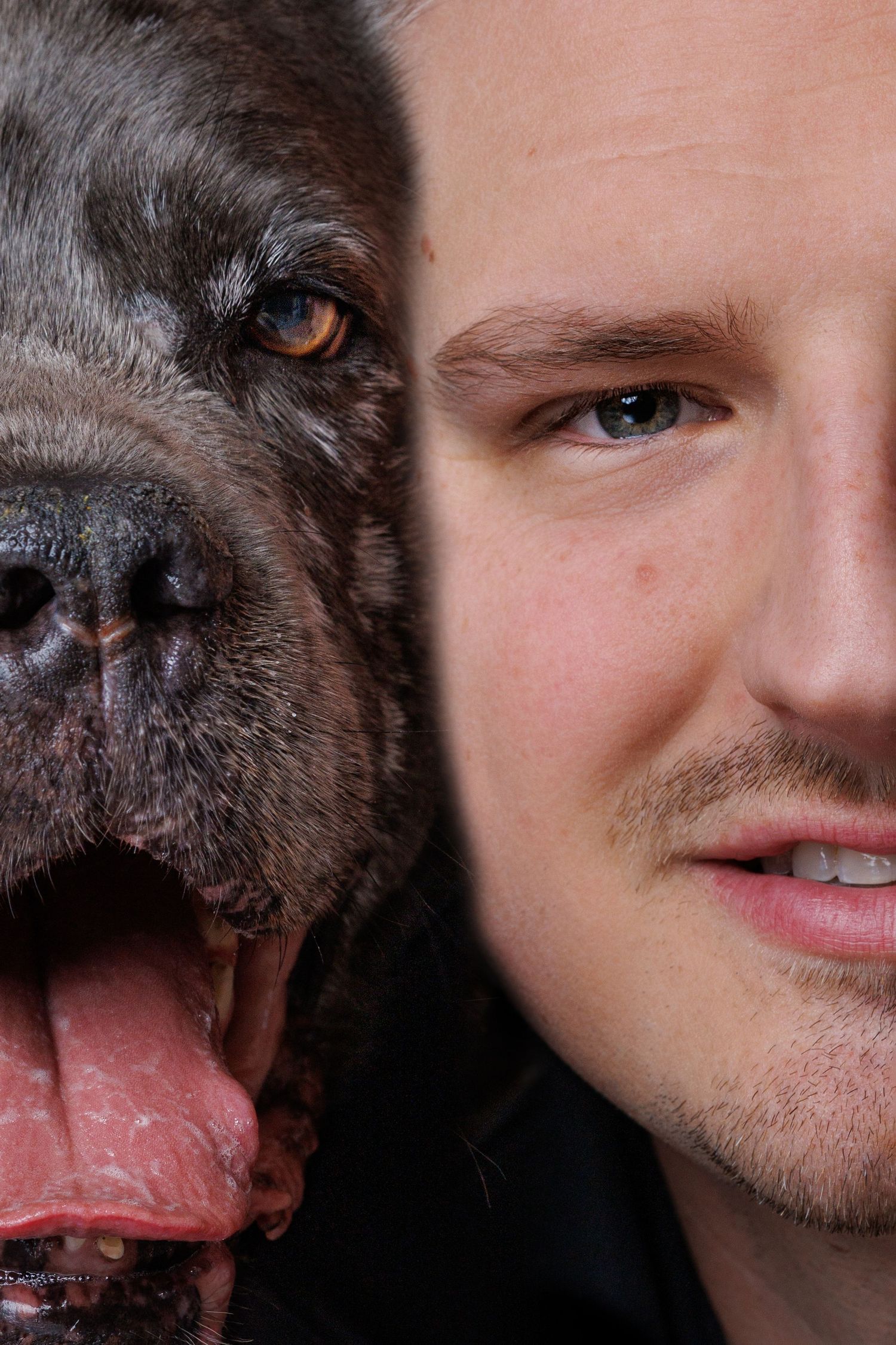 Close-up portrait of a young man with light stubble cheek-to-cheek with a dark-coated dog with tongue out.