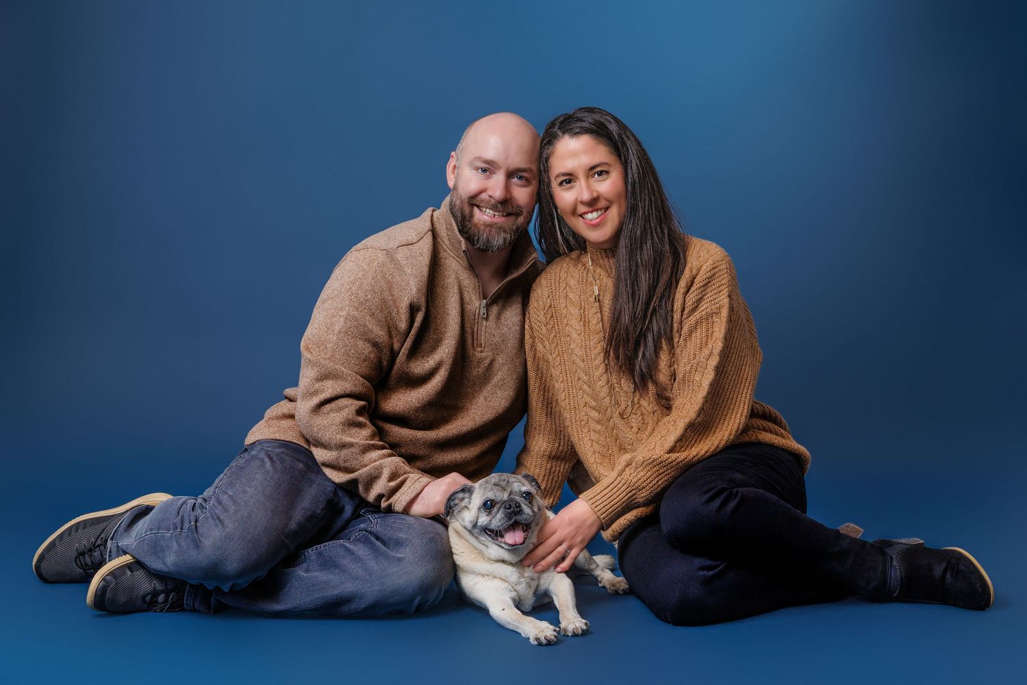 Smiling couple sitting together with their pug dog on blue studio background.