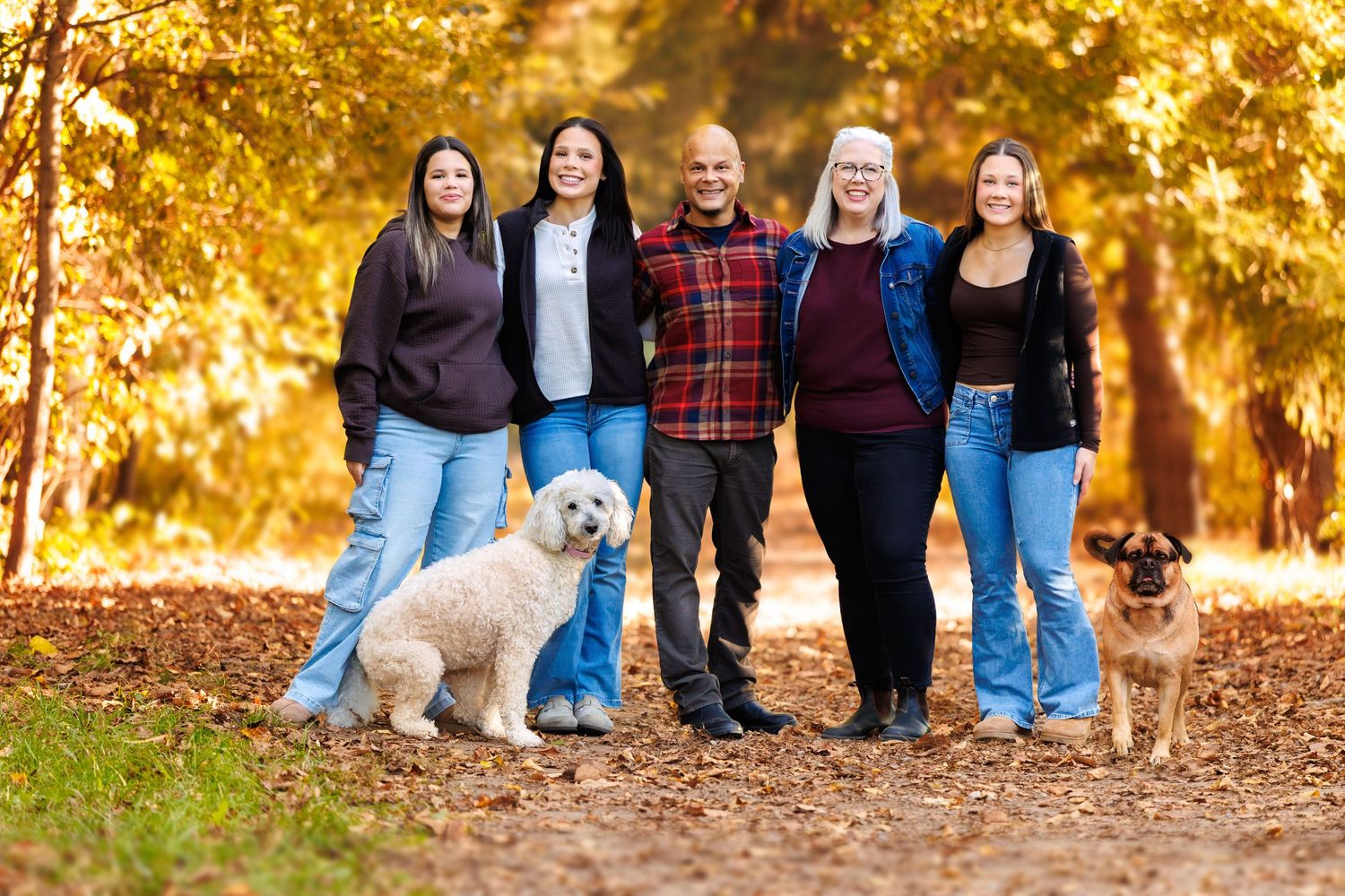 Family of five poses with two dogs on a leaf-covered path surrounded by golden autumn trees.