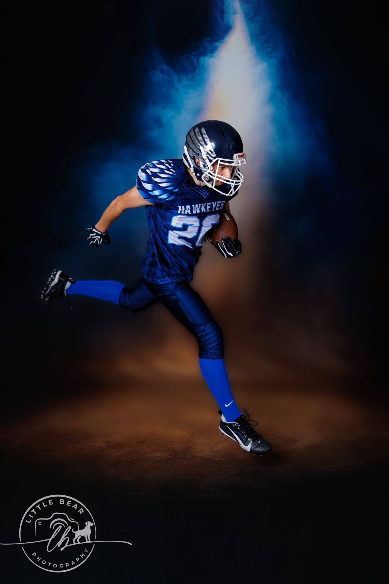 Youth football player in blue Hawkeyes uniform running with ball in dramatic studio portrait.
