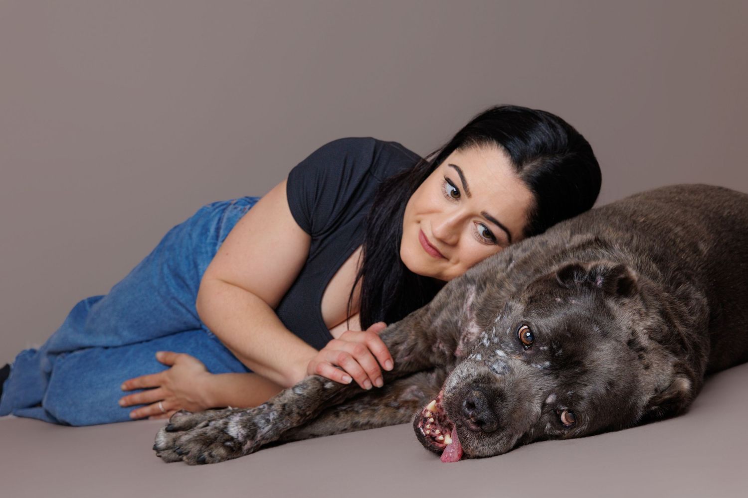 Woman with dark hair lying beside a large gray dog, both relaxed on the floor in a studio portrait.