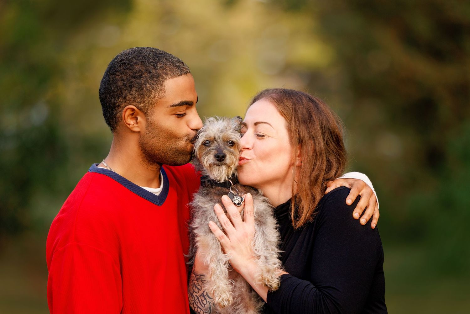 A couple kisses a small gray terrier dog on each cheek in an outdoor park setting.