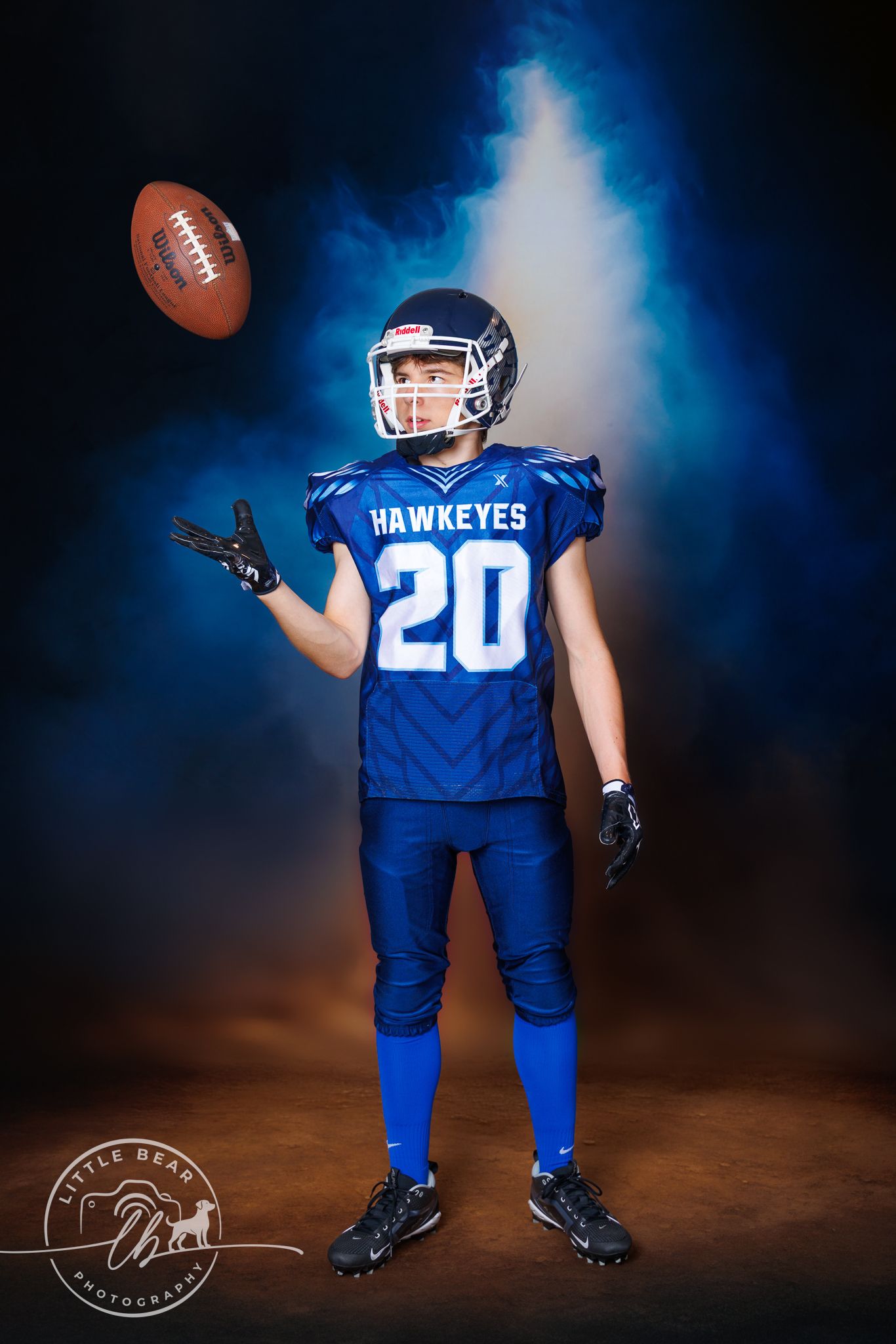 Young football player in blue Hawkeyes #20 jersey tosses a football in a dramatic studio portrait.