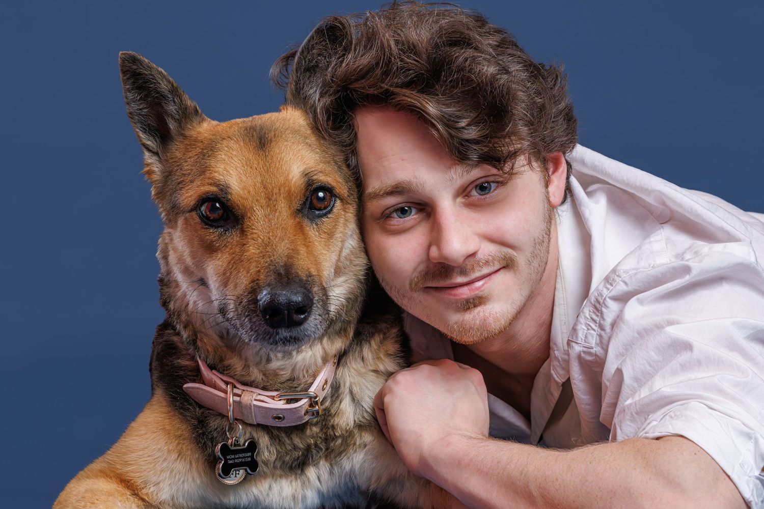 Young man smiling and hugging a German Shepherd mix dog against a blue studio background.