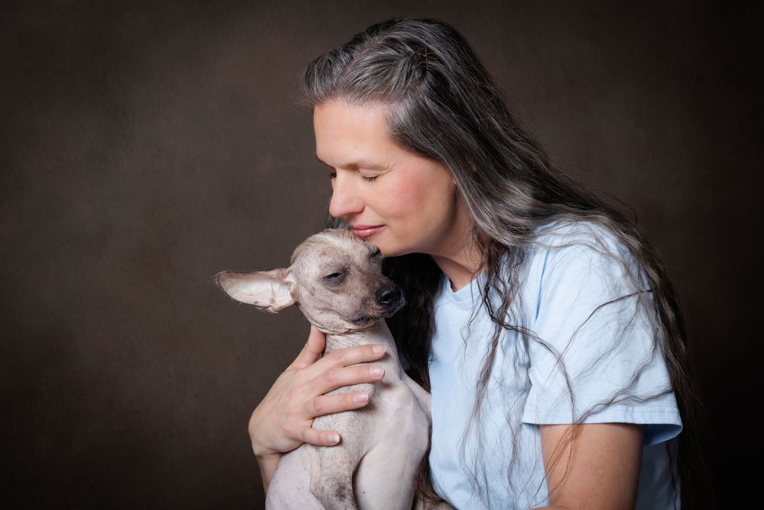 A woman with long gray hair tenderly holds and nuzzles a small Chihuahua dog against a dark studio background.