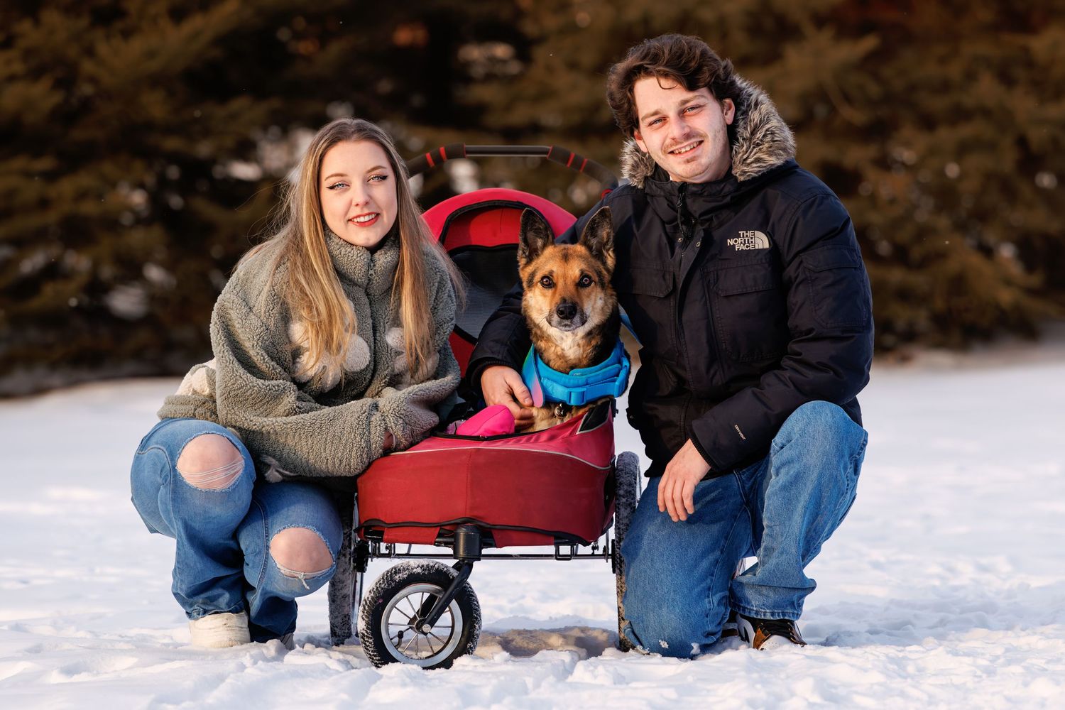 Two people kneeling in snow with a dog sitting in a red wagon, surrounded by snowy trees in winter.