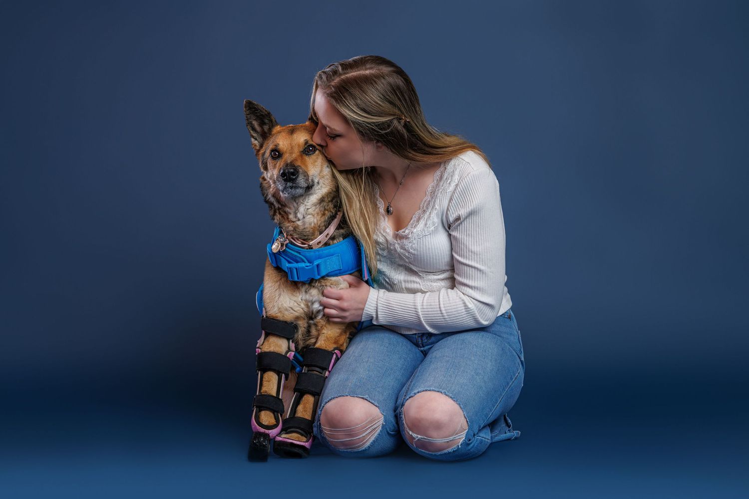 Woman in white top kissing her dog wearing a blue orthopedic leg brace on a blue studio background.