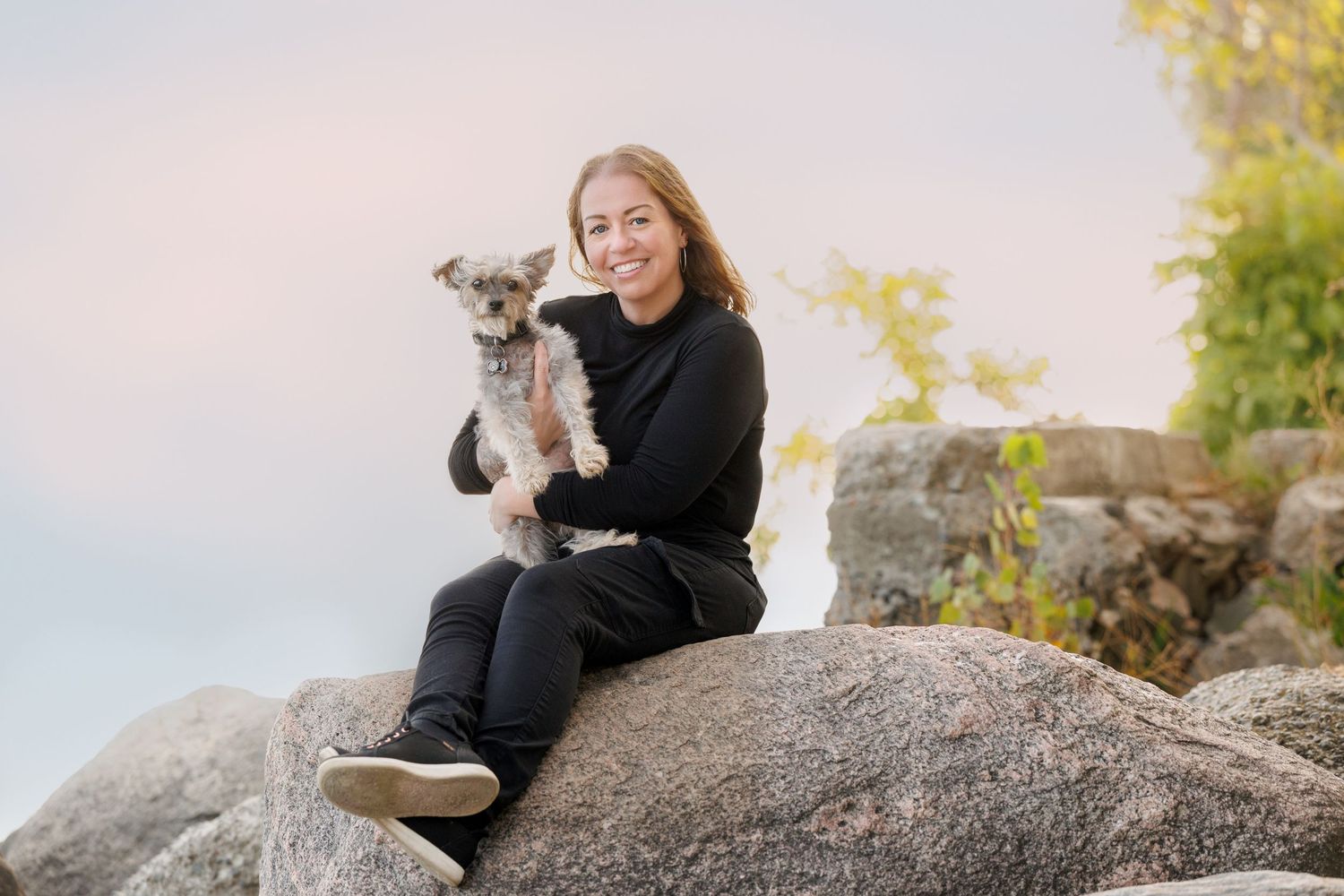 A smiling woman in black sits on a large rock holding a small gray Yorkshire Terrier dog.