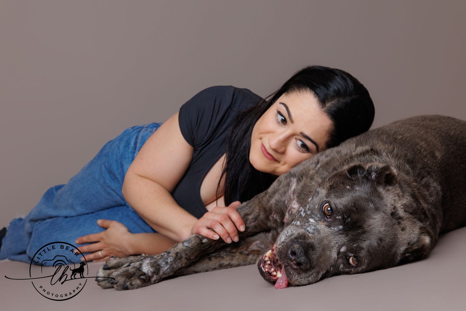 Woman with dark hair lying beside a large brindle Cane Corso dog in a professional studio portrait.