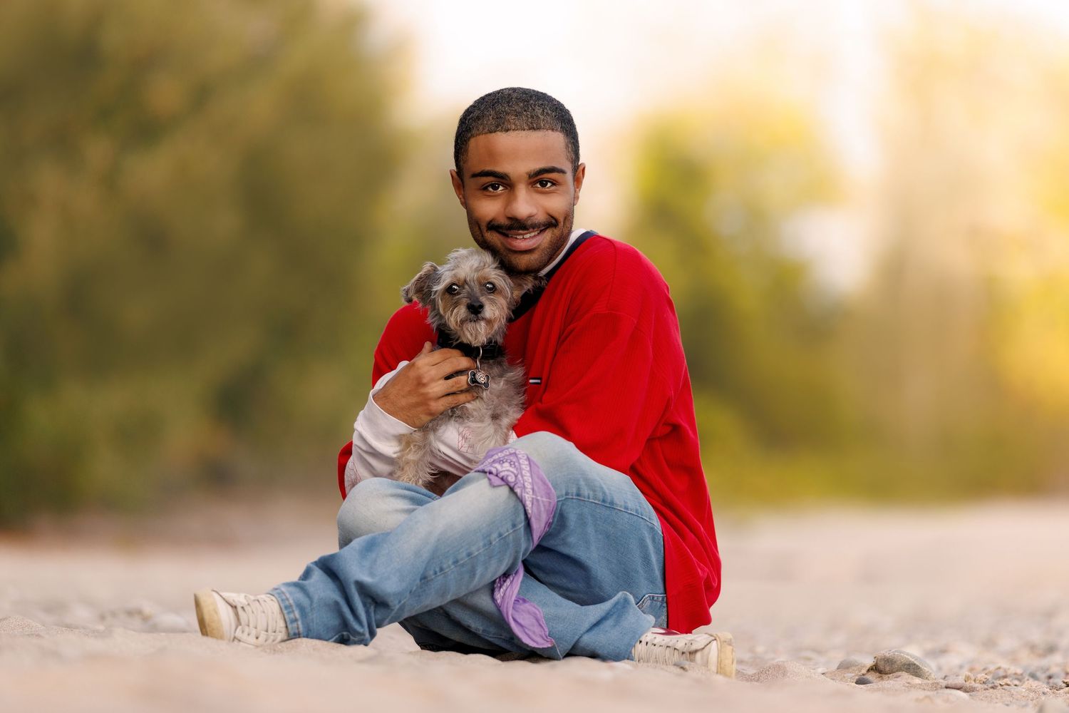 A smiling man in a red hoodie sits on sand hugging a small gray Yorkshire Terrier dog.
