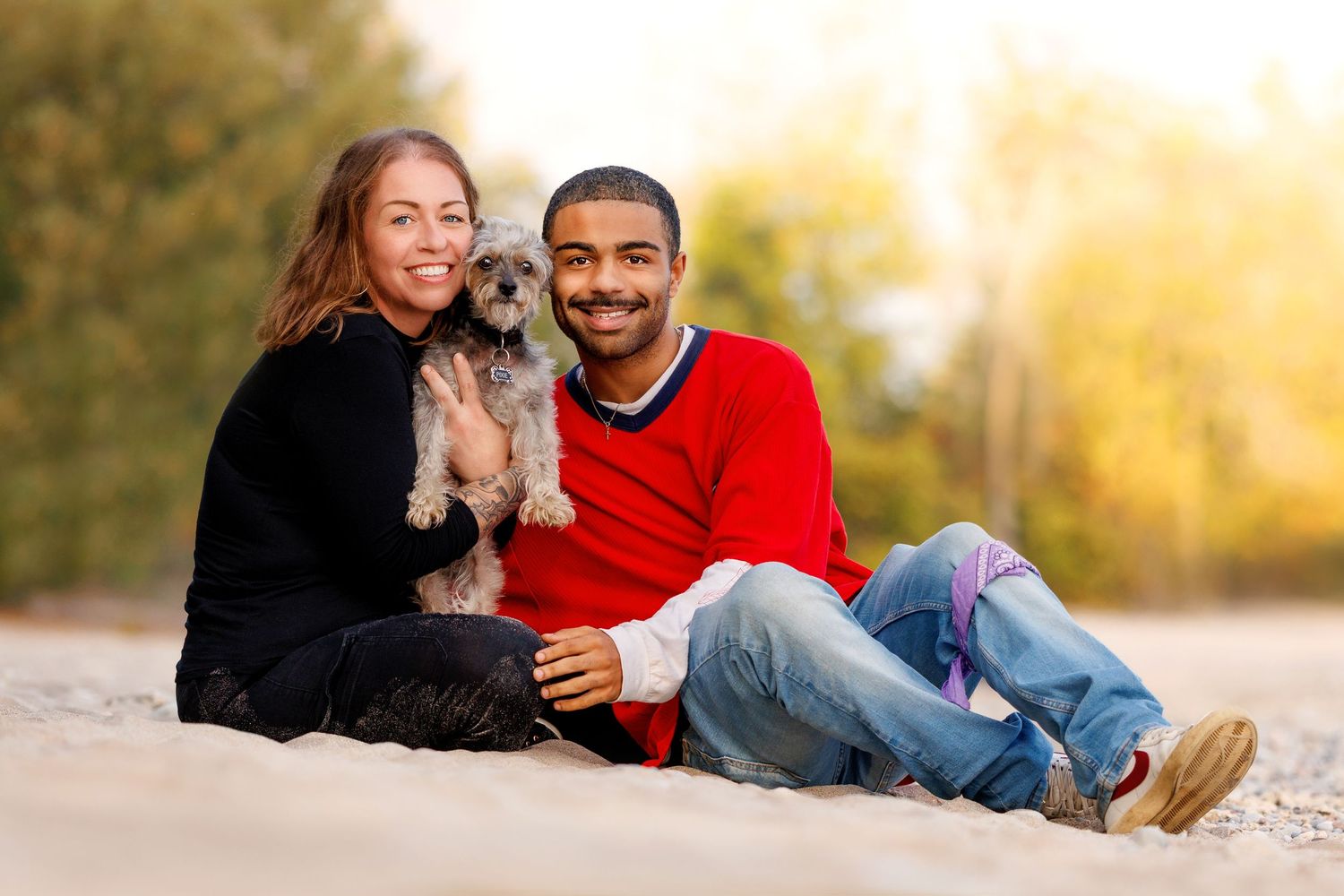 A smiling couple sits on sandy ground outdoors holding a small gray Yorkshire Terrier dog.