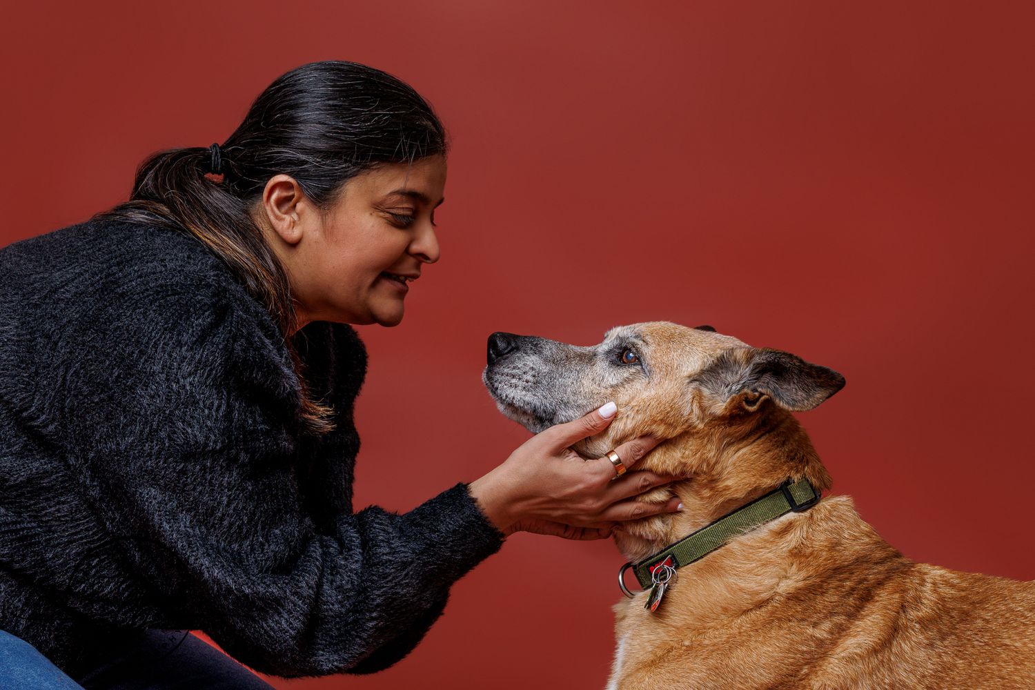 Woman in black jacket tenderly holds the face of a golden brown dog against a red background.