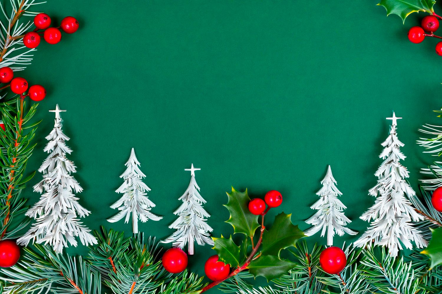 Green Christmas background with white paper trees, holly leaves, red berries, and pine branches forming a festive holiday border frame.