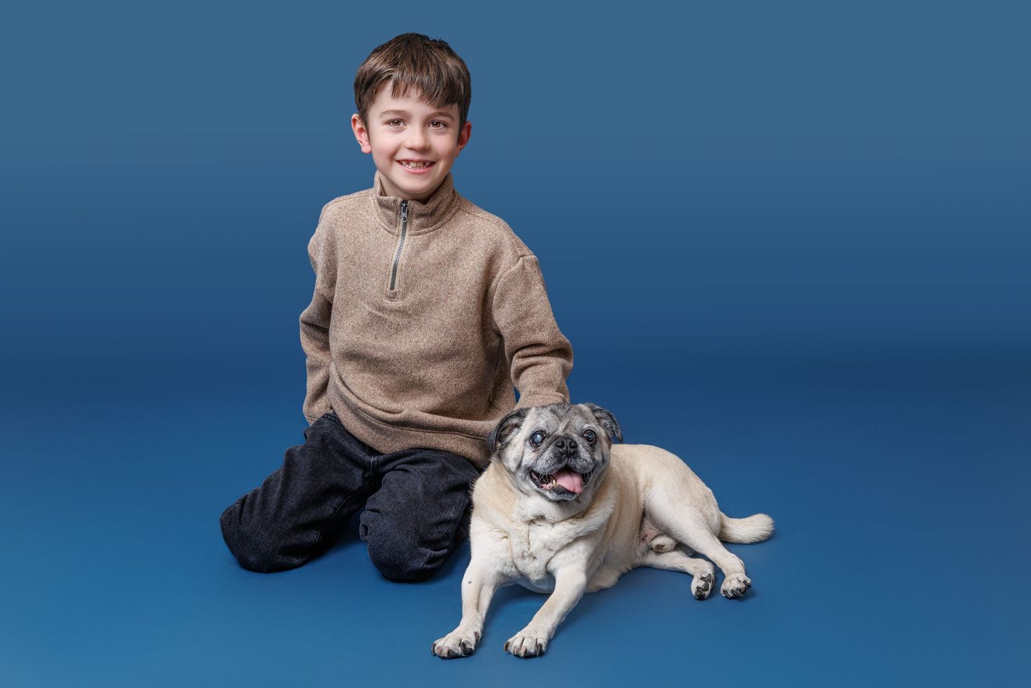 A smiling boy in a beige sweater sits on a blue background with a friendly pug dog beside him.
