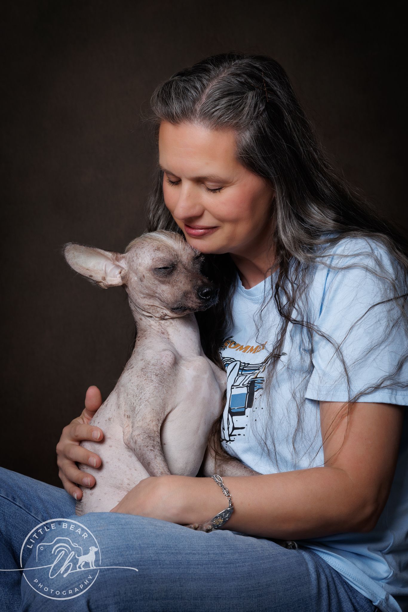 Woman in blue t-shirt tenderly holds a hairless Chihuahua dog in a studio portrait photo.