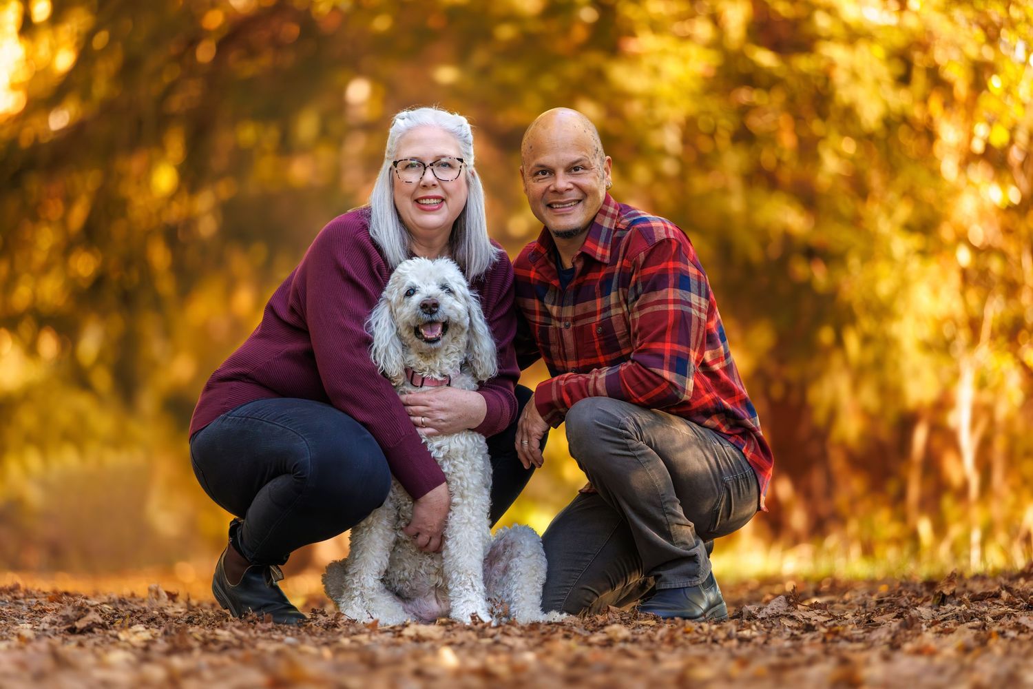 Couple smiling with white dog outdoors in autumn forest with golden leaves, wearing casual fall clothing.