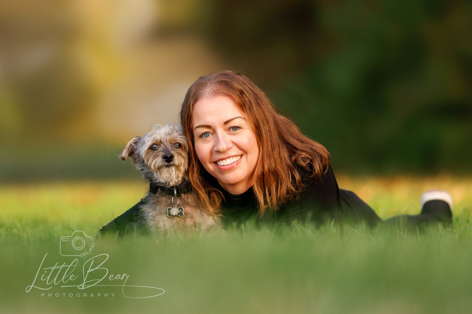 Woman and small terrier dog lying in green grass, smiling outdoors in a park during golden hour.