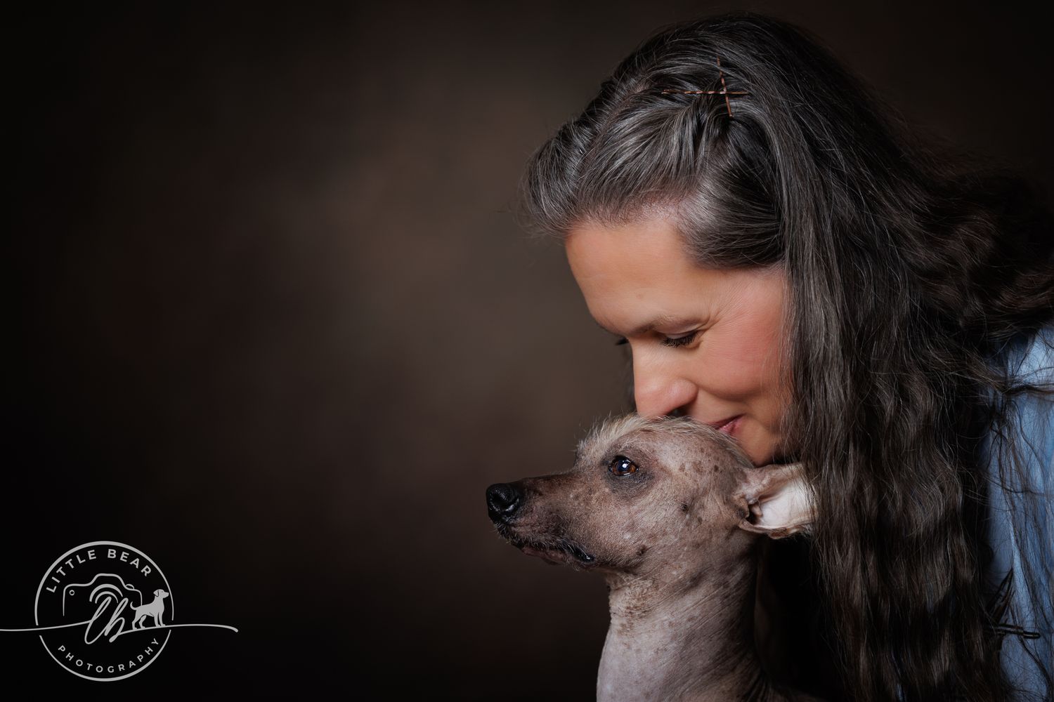 A woman tenderly kisses a hairless dog in a warm studio portrait photo.