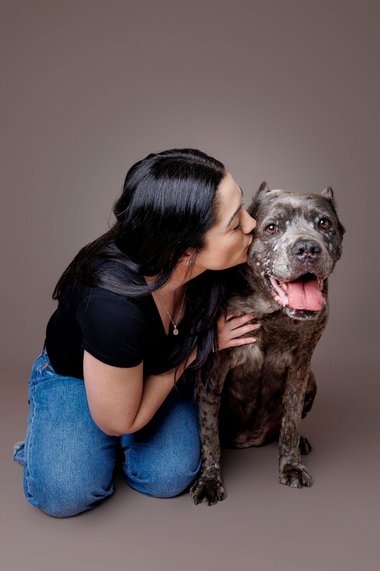 Woman in black top and jeans kissing her brindle pit bull dog on the cheek in a studio portrait.