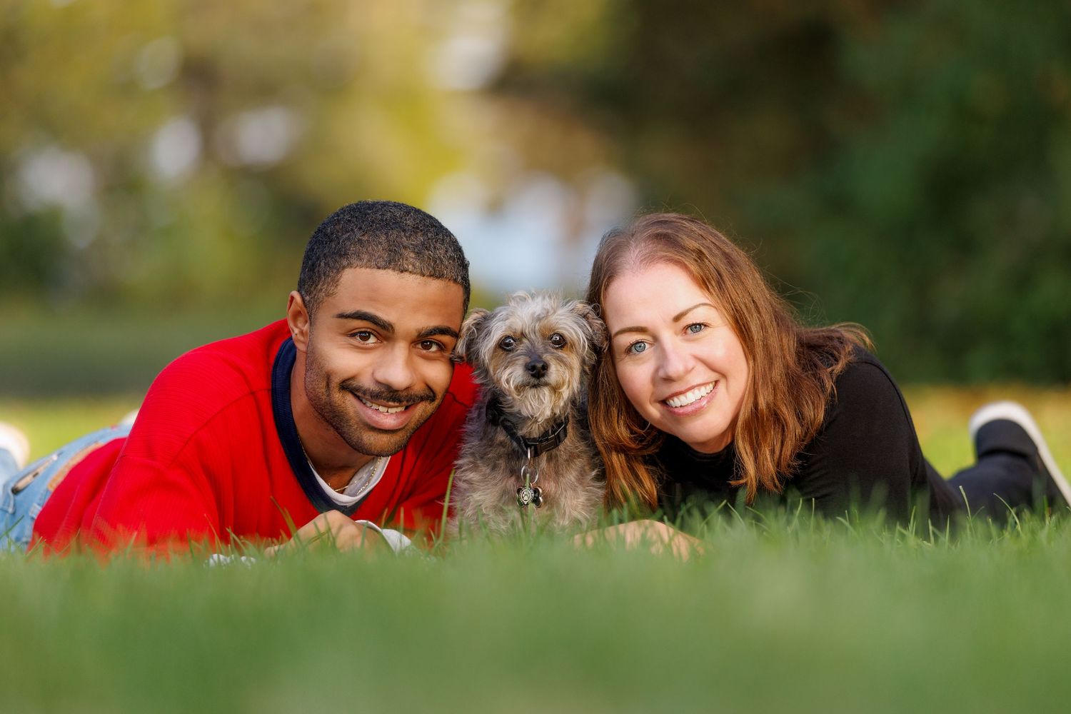 A smiling couple lies on green grass with their small gray terrier dog between them.