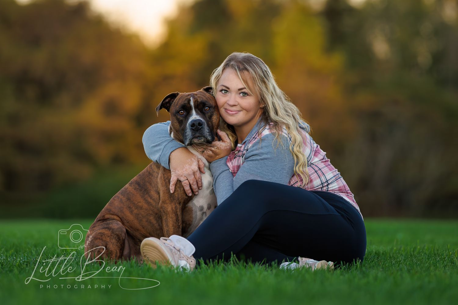 A smiling blonde woman sits on green grass hugging her brindle Boxer dog with autumn foliage behind.
