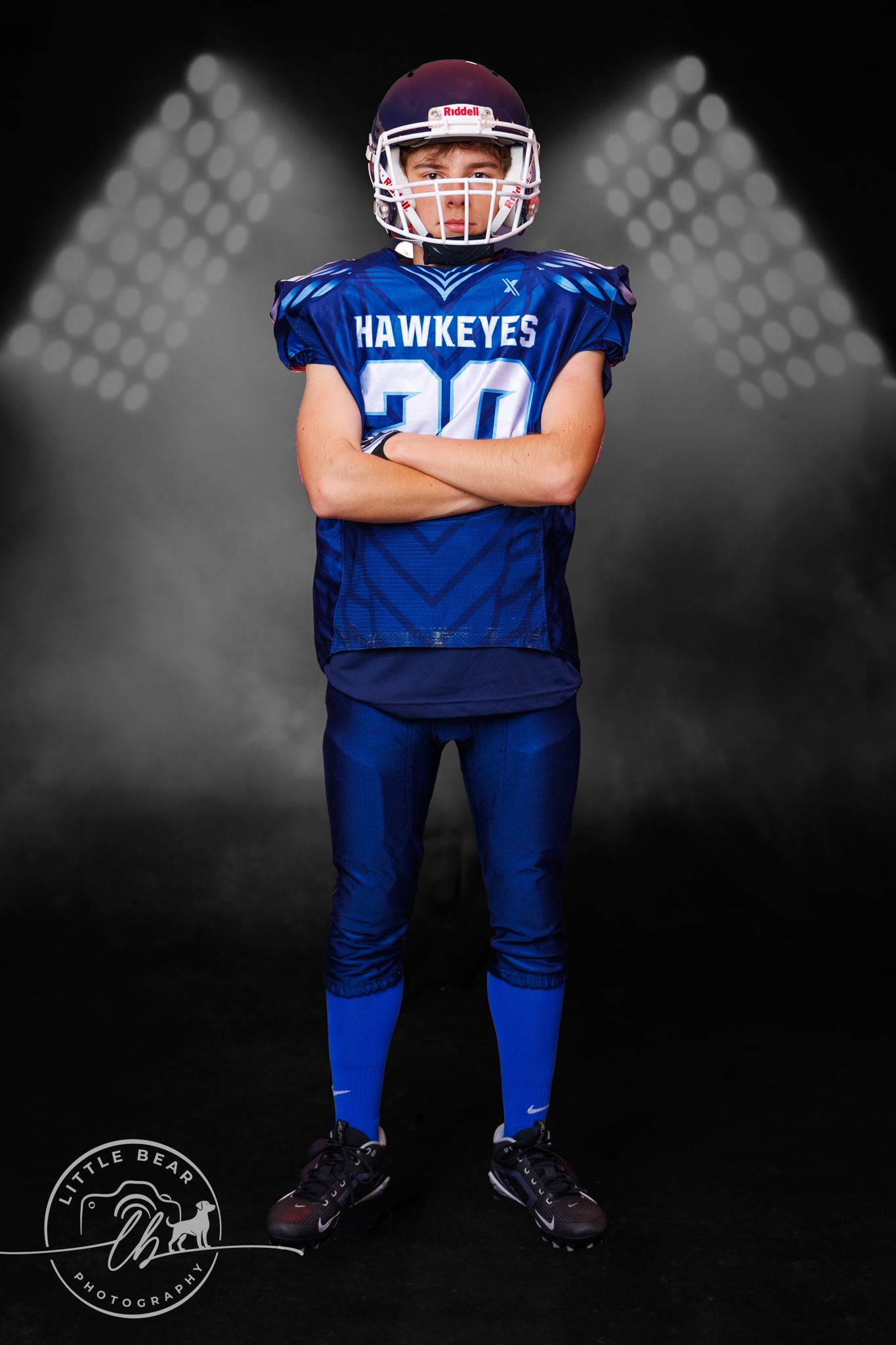 Young football player in blue Hawkeyes jersey number 20 poses with arms crossed under stadium lights.