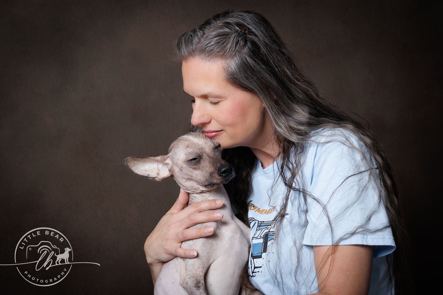 Woman with long gray hair tenderly holding a small hairless Chihuahua dog in a studio portrait.