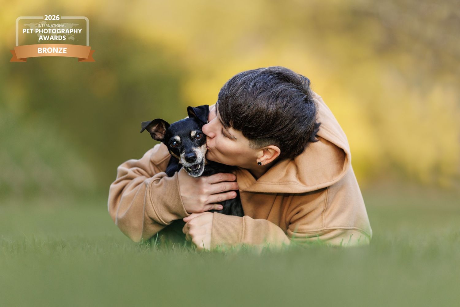 Person in beige hoodie kissing small black dog while lying in green grass, award-winning pet photography.