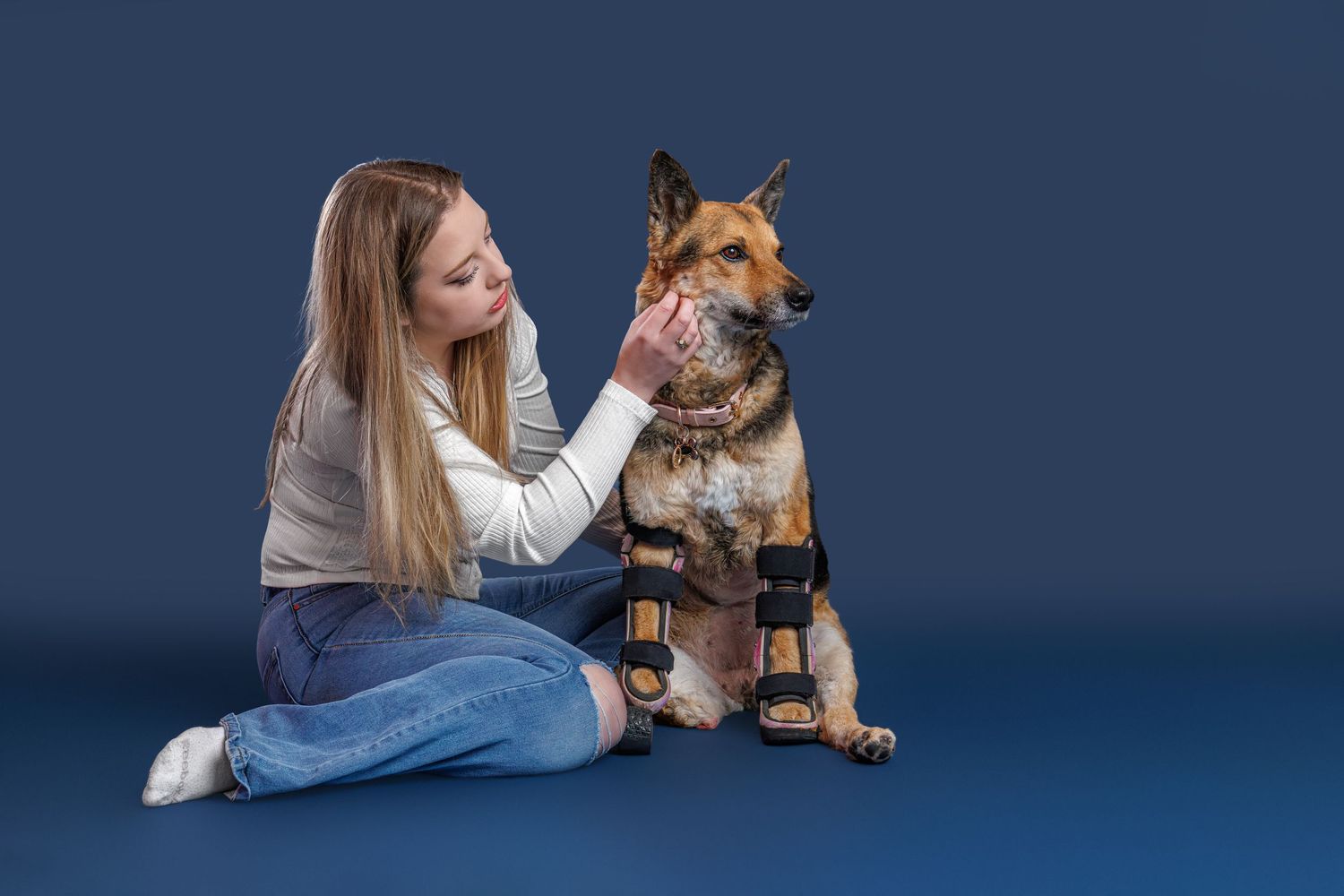 Woman sitting on blue floor petting a German Shepherd dog wearing orthopedic leg braces.