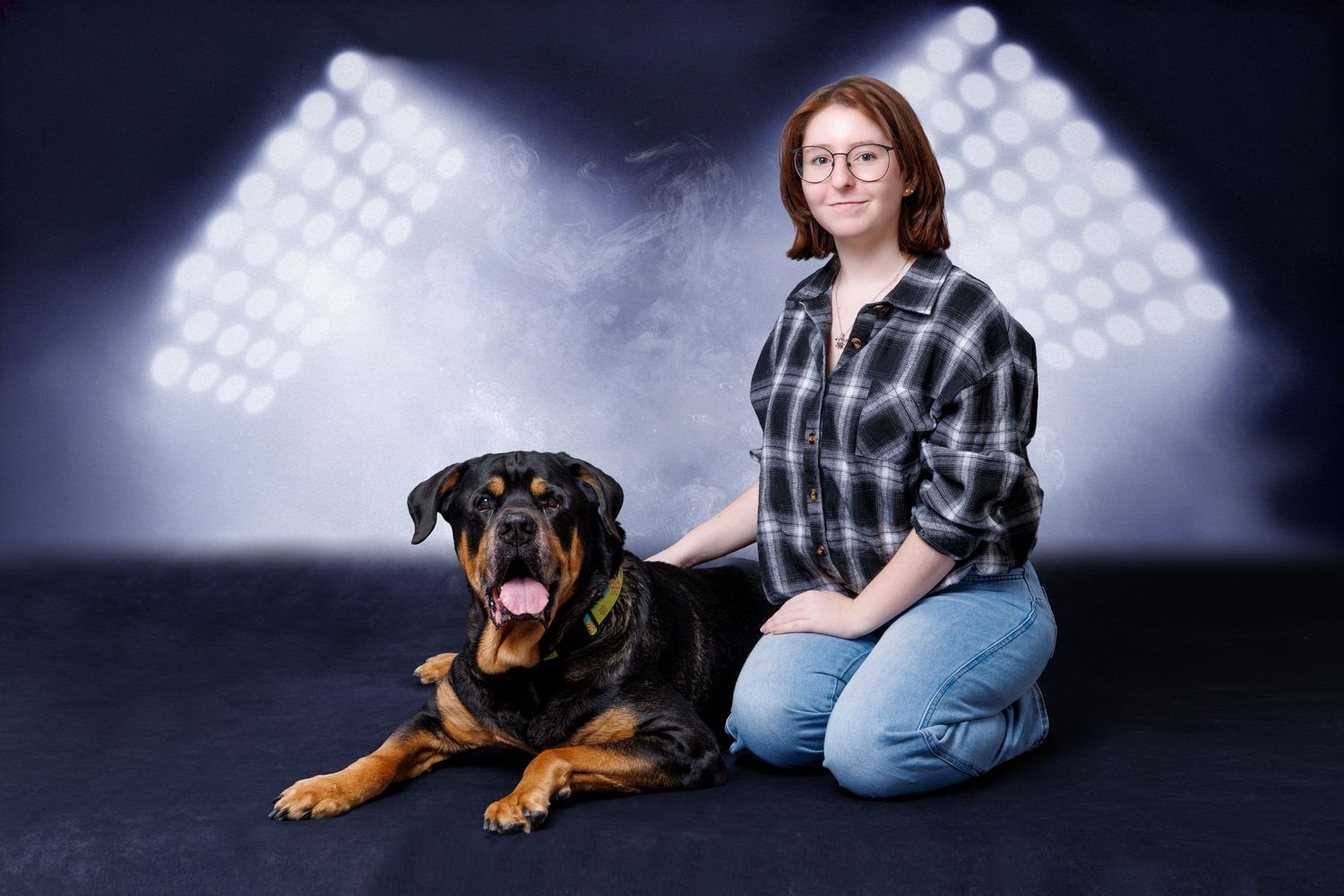 Young woman with glasses sits beside a Rottweiler dog against a dramatic studio backdrop with spotlights.