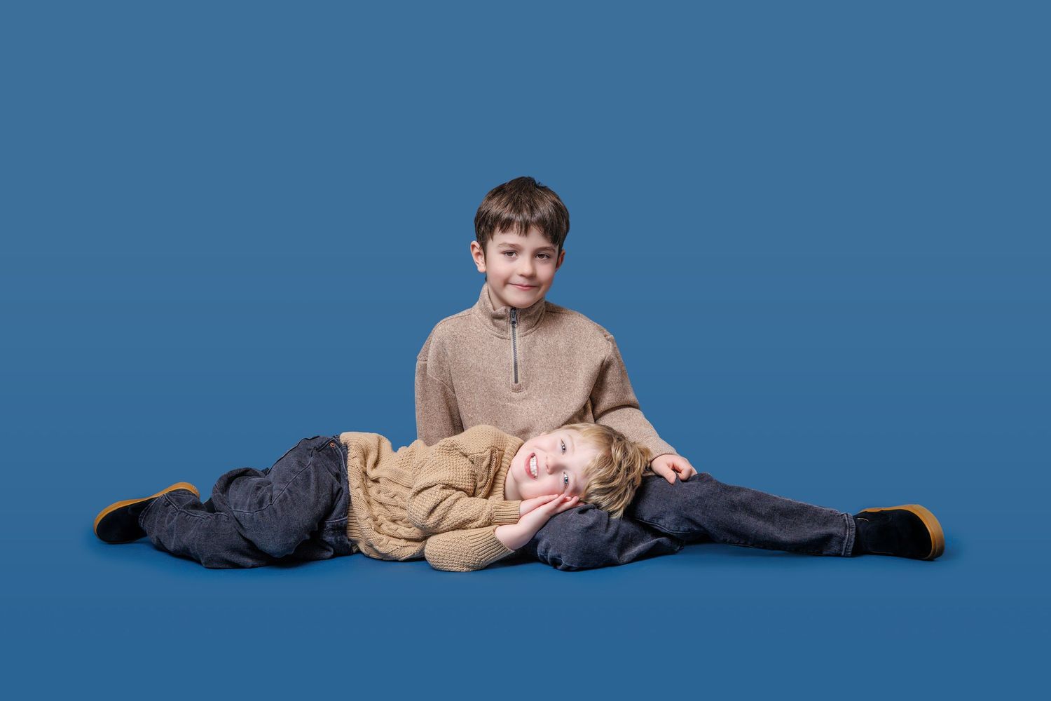 Two boys posing together on blue background, one sitting up and one lying down smiling.