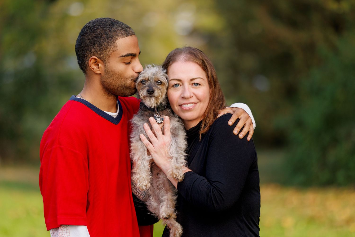 A young man kisses a small gray terrier as a smiling woman holds the dog outdoors.